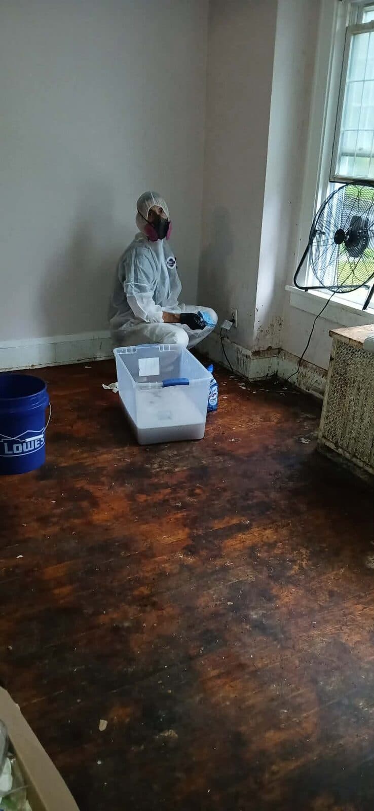 Crew member in a protective suit and respirator kneeling beside a bin of cleaning supplies on a stained floor